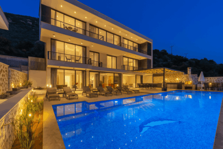 Villa Falcon Royal in Kalkan with private infinity pool, modern three-story design, luxury sun loungers, and illuminated evening ambiance surrounded by nature.